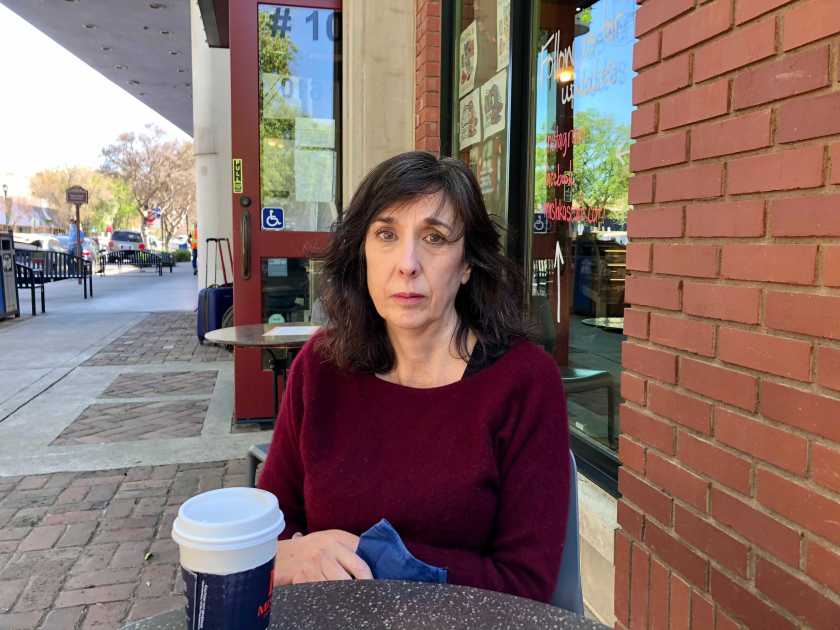 A woman sits at a table outside a brick storefront with a to-go cup of coffee