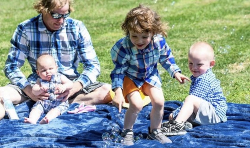 A man holds an infant while two young kids play with bubbles on a blanket outside.