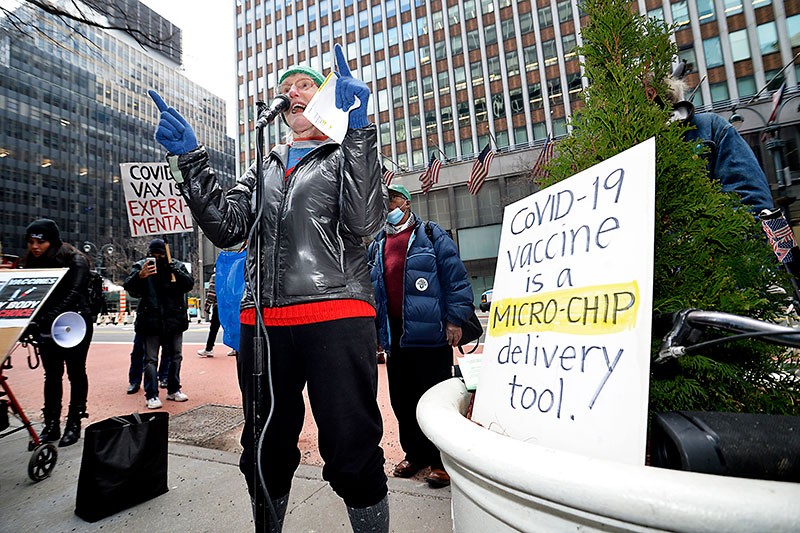 A woman talks into a microphone, surrounded by people holding various anti-vaccine placards.