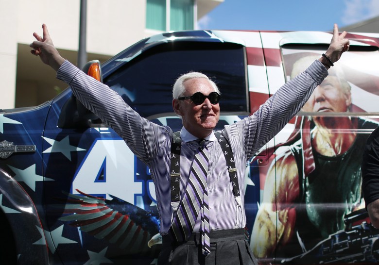 Roger Stone stands with his arms raised wide, flashing two V-shaped hand signs, a pose made famous by former President Richard Nixon. He stands in front of a large vehicle with an American flag design and an image of a gun-toting action star with former President Donald Trump’s face superimposed onto it.