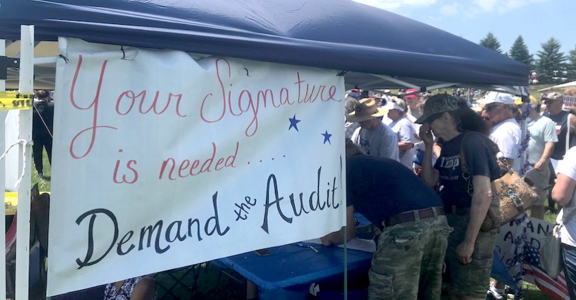 A booth at a rally with a sign hanging from it’s covering that reads “Your signature is needed.... Demand the Audit!”