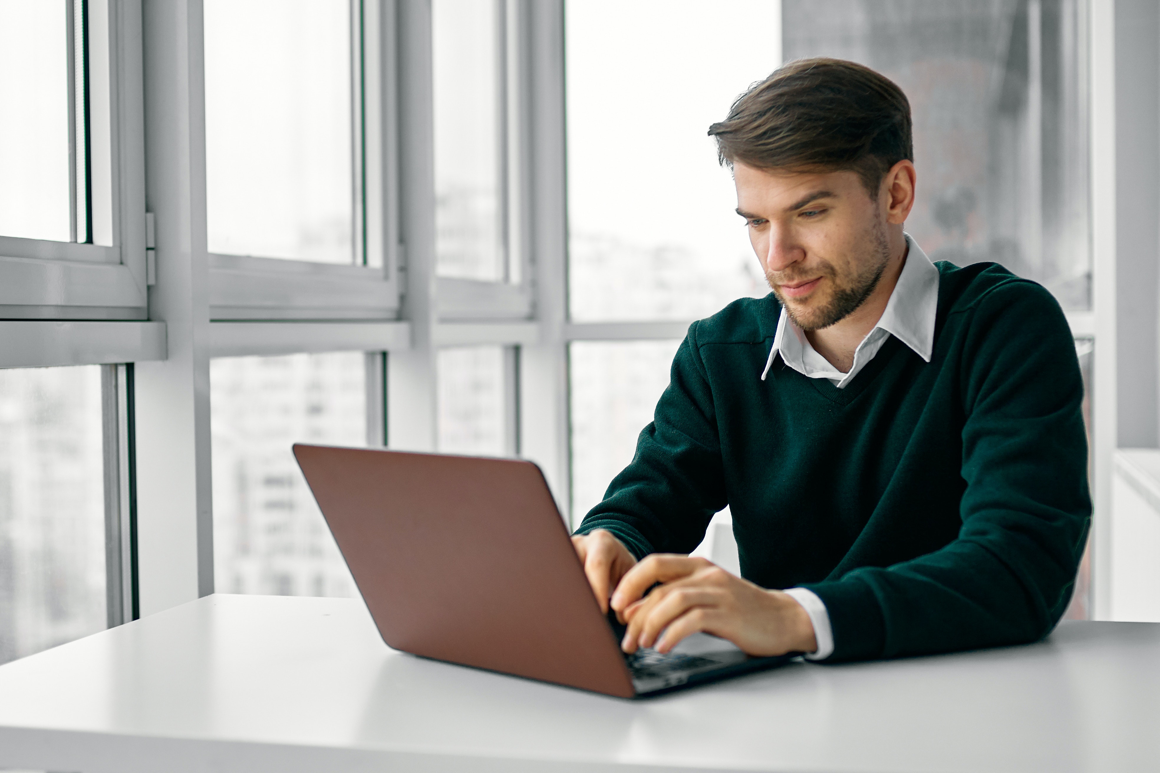 A man wearing a green sweater sits at a white table typing on his laptop.