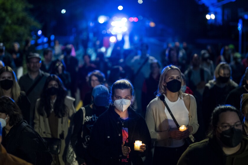 a crowd of mostly women holding candles stand together on a darkened street with lights in the background