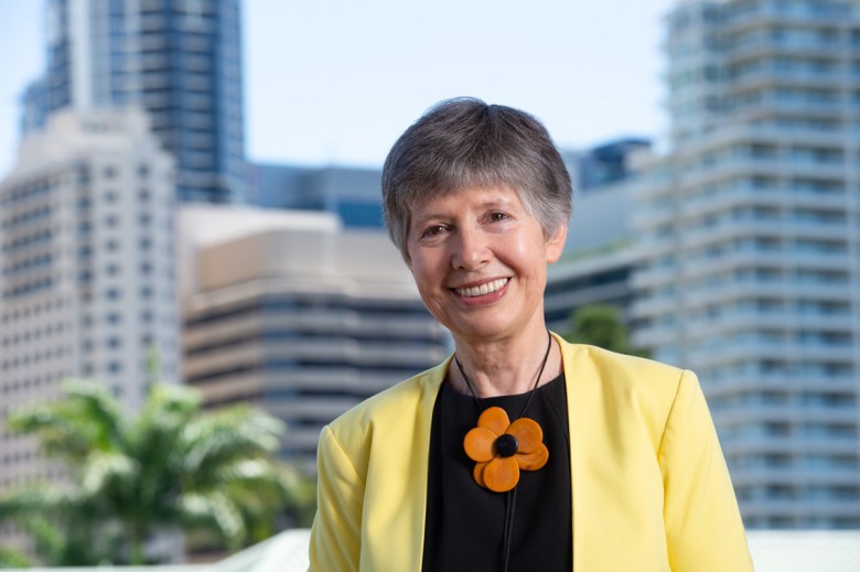 A gray-haired smiling woman wearing a yellow jacket and a bolo necklace with a large orange flower over a black top poses in front of a blurry city background.