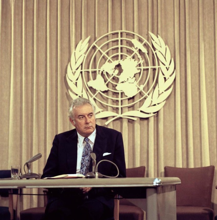 Colour photo of middle-aged man in suit sitting behind desk with UN symbol in background. - click to view larger image