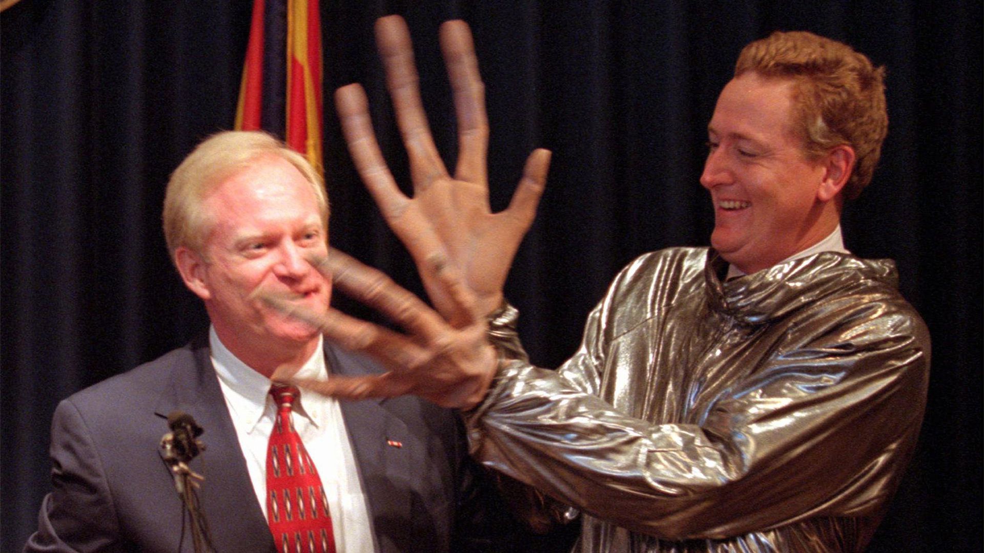 A man in an alien costume stands in front of a man at a podium.