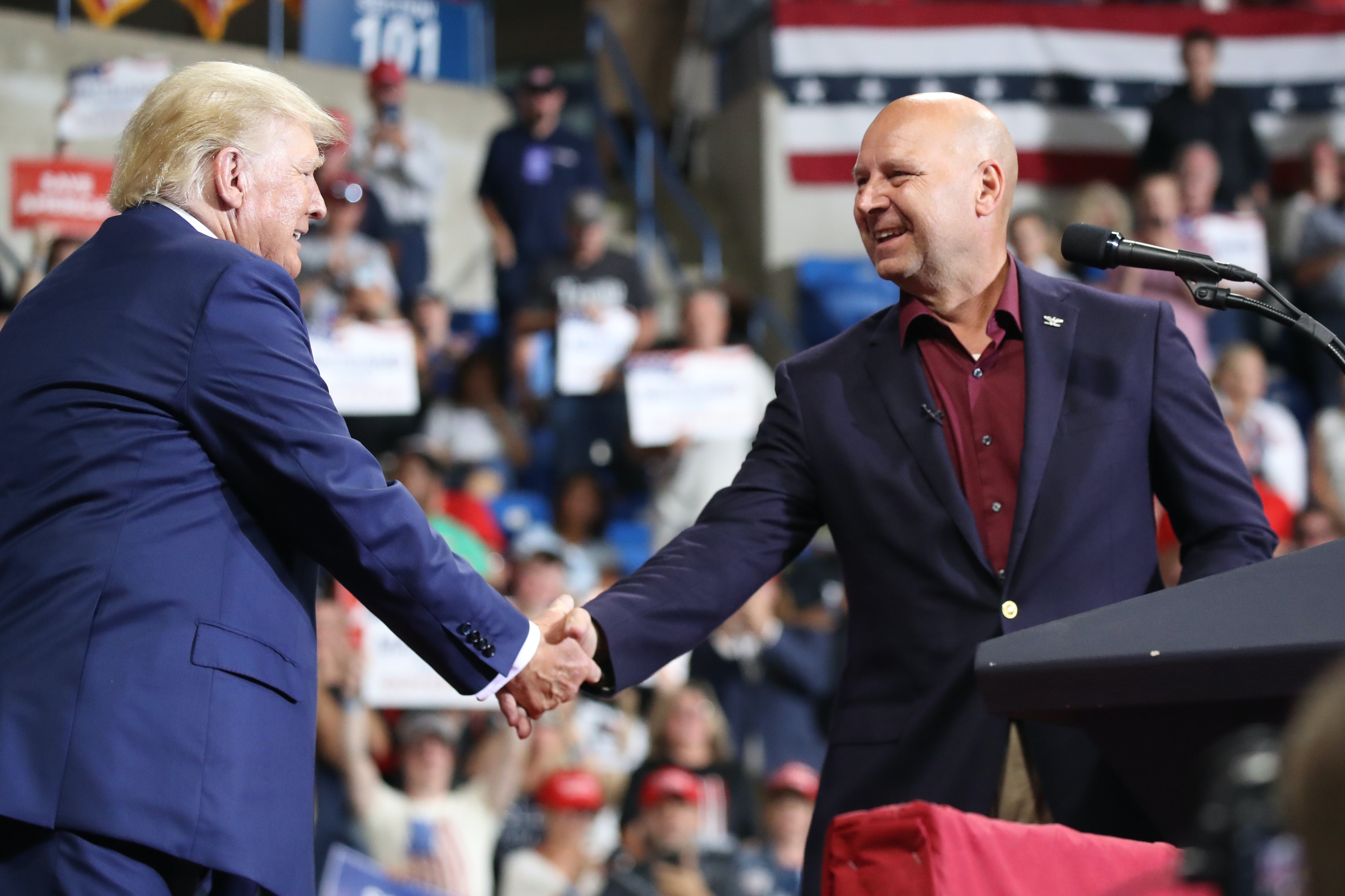 Two middle-aged white men shake hands on a stage in front of a large crowd.