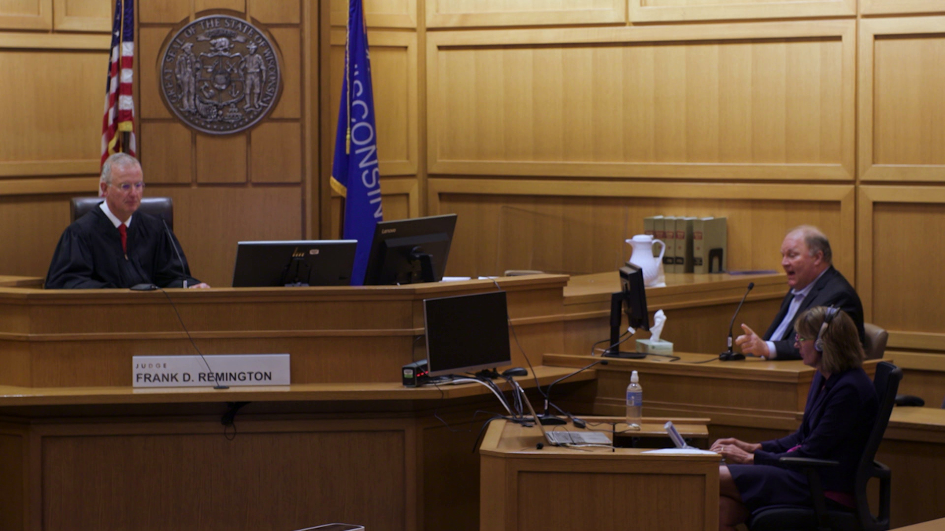 Michael Gableman is seated in the witness box of courtroom with wood-paneled walls and furniture, with a judge seated at a dais, facing two screens and in front of the U.S. and Wisconsin flags and the seal of the state of Wisconsin, and with a court reporter wearing headphones seated at a desk below.