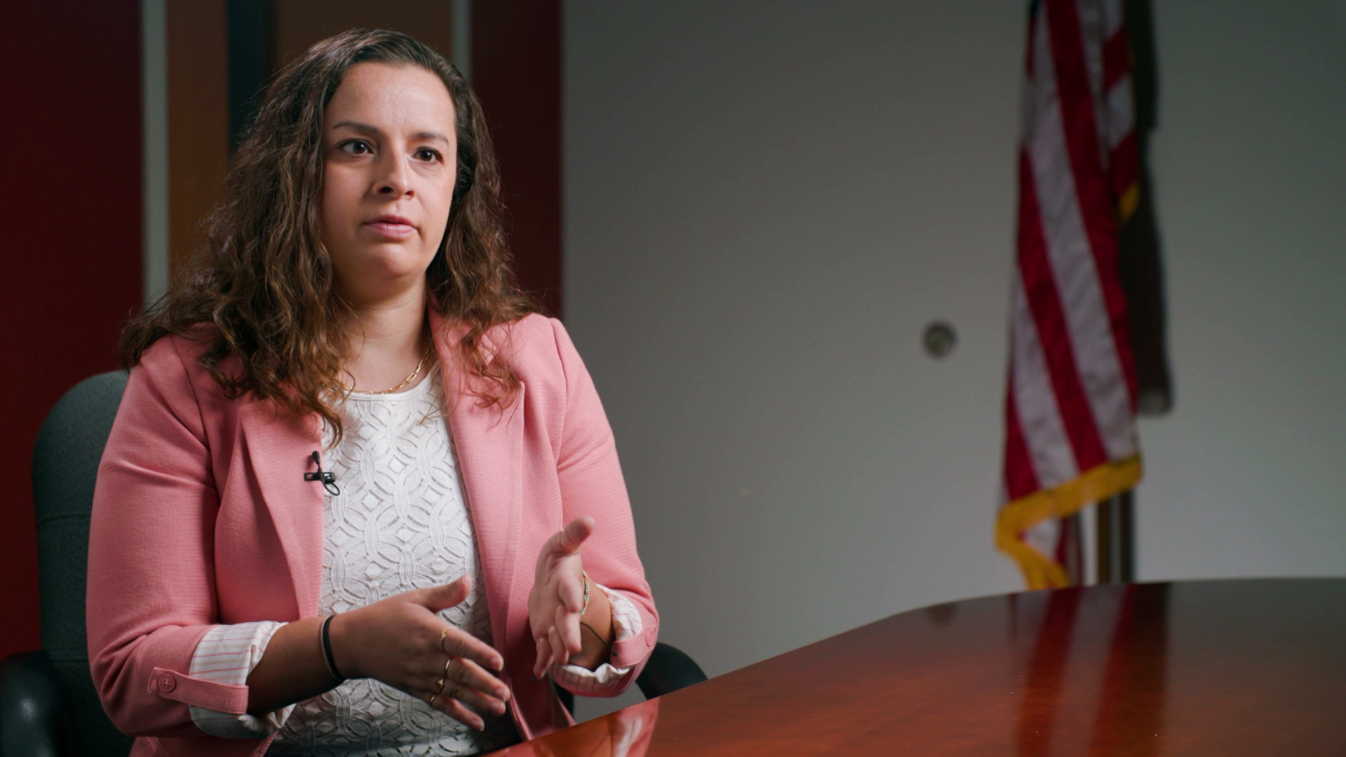 Rachel Rodriguez sits at a table and gestures with her hands, with a door and U.S. flag in the background.