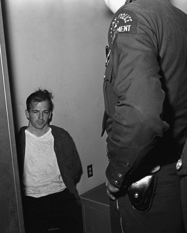 Lee Harvey Oswald sitting, while handcuffed, with a police officer standing over him in a doorway.