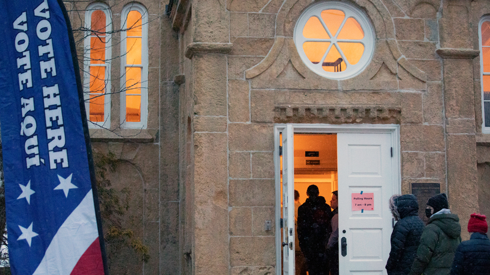 People stand in a line outside a double doorway to a stone masonry building with arched and roundel windows, with a vertical banner reading "Vote Here" and "Vote Aquí" standing next to a tree at its corner.