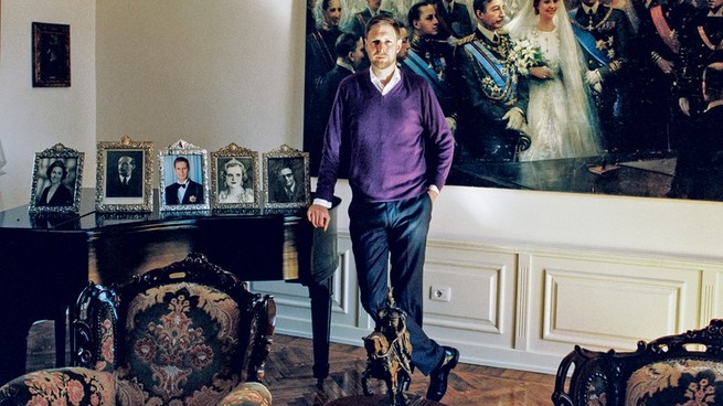 photo of a man in purple sweater standing in front of a large painting of a royal wedding and beside a piano on which five portrait photos are displayed