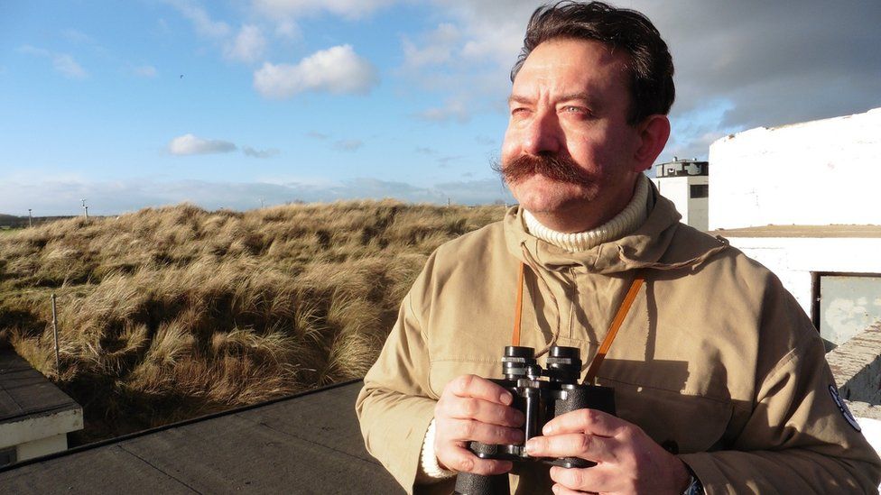 Neil Storey sports a magnificent moustache and holds binoculars