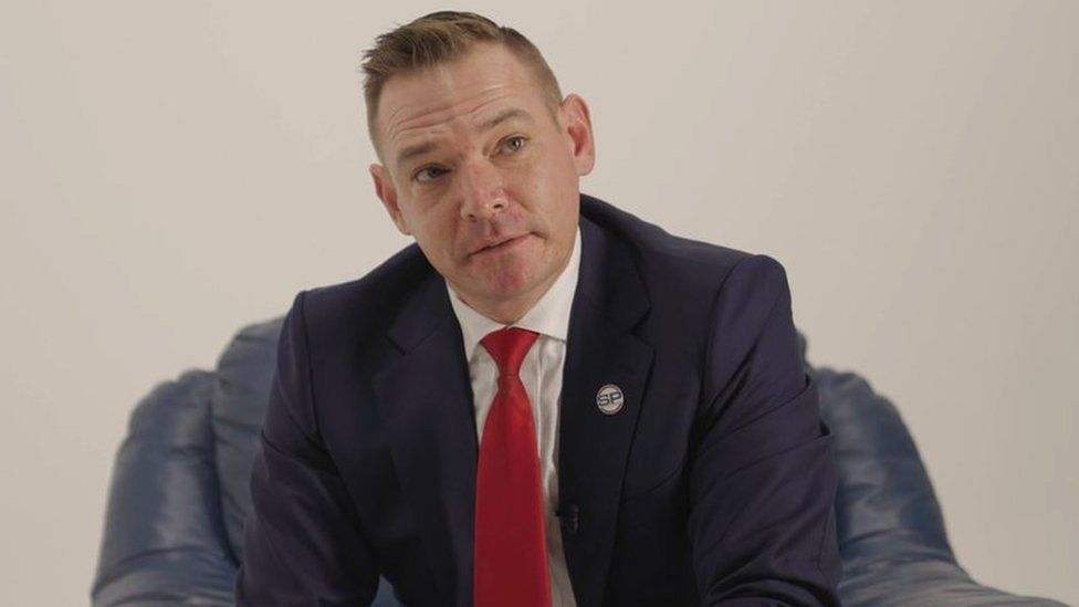 White man in suit and tie sitting on an armchair with a SP badge on his lapel