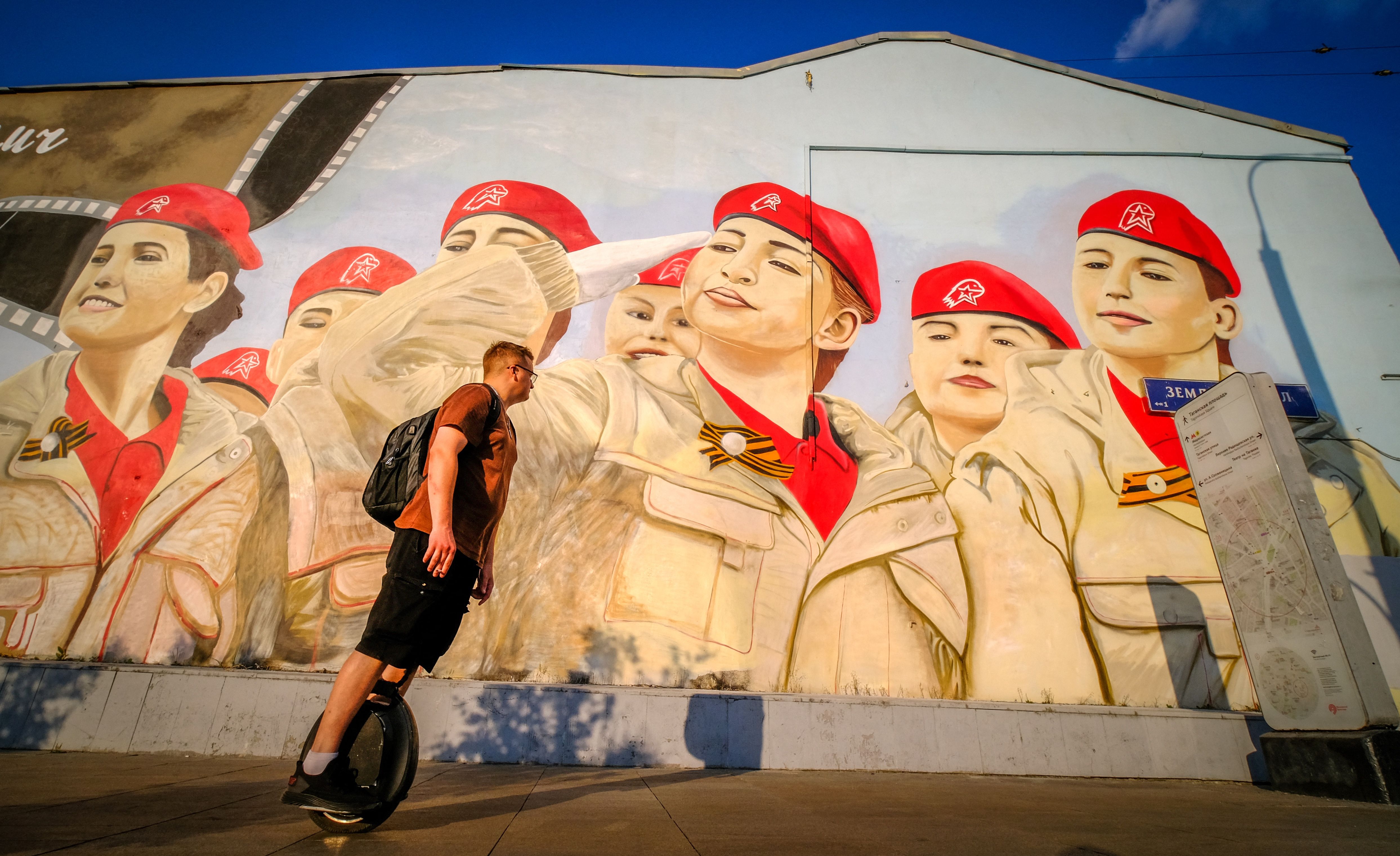 A man rides a unicycle past a house decorated with a mural that depicts members of the Russian Patriotic Youth movement Yunarmiya in Moscow in 2022. The painted children wear red berets and tan uniforms. One has her hand to her forehead in a military salute.