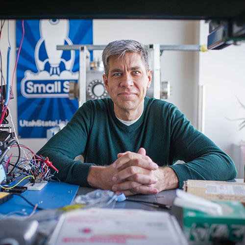 Photo: ENG professor of electrical and computer engineering, Josh Semeter poses for photo in his lab on Wednesday, January 20, 2016. A white man with silver grey hair sits with hands folded in front of him and resting on a desk cluttered with papers, wires, and various hardware constructs. He wears a dark green sweater. A lab filled with random mechanics is shown behind him as a large blue and white spaceship banner hangs on the wall behind him.