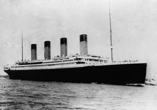 A black-and-white view of the Titanic on the water. There are smaller boats near it. 