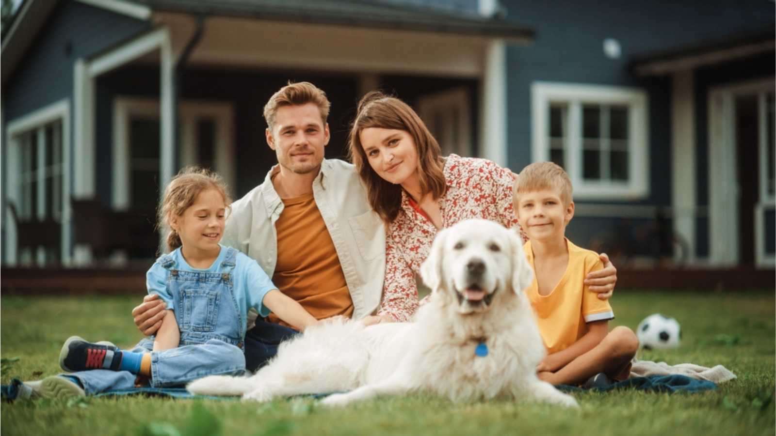 Portrait of a Happy Young Family Couple with a Son and Daughter, and a Noble White Golden Retriever Dog Sitting on a Grass in Their Front Yard at Home. Cheerful People Looking at Camera and Smiling.