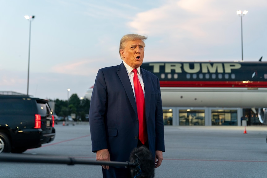 Donald Trump, in suit, speaks with reporters in front of a plane that has his name on it.