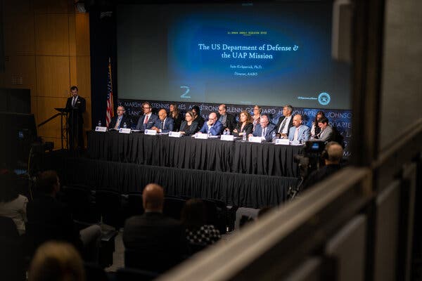 Several members of NASA's unidentified anomalous phenomena independent study team sit at a long table on a stage at NASA headquarters with a large screen behind them that reads "The US Department of Defense & the UAP Mission."