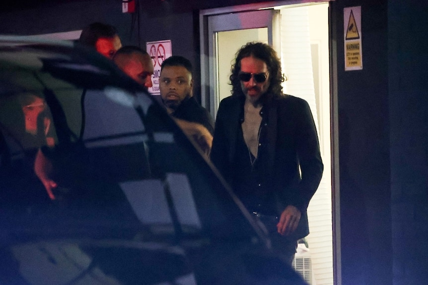 A man wearing sunglasses can be seen walking out a door, surrounded by security guards