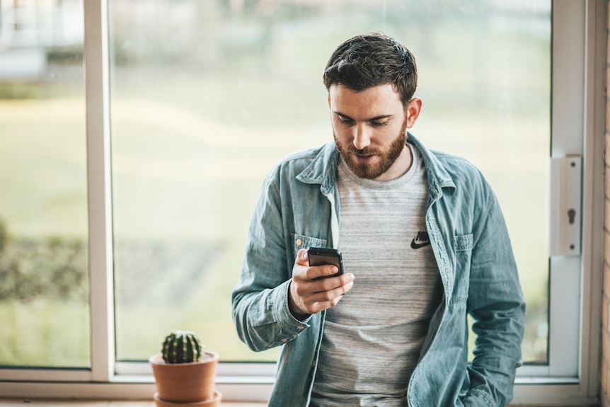 A young Caucasian man with brown hair and a short beard leans against a windowsill and looks intently at his phone. 