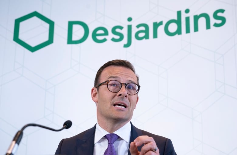 Guy Cormier has dark hair, round glasses and is wearing a suit with a blue tie. He is speaking into a small microphone and behind him it reads: Desjardins.