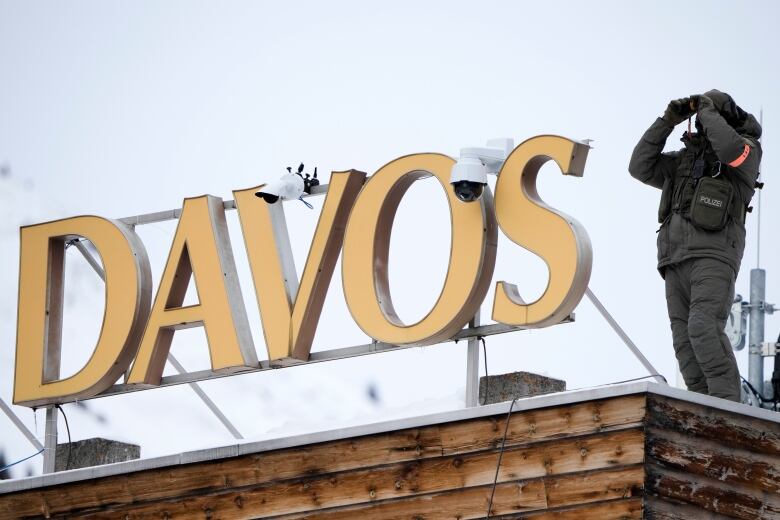 A police officer clad in grey holds up binoculars to his eyes as he stands beside a sign that says Davos.