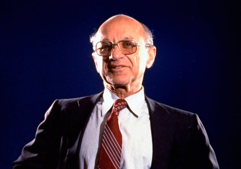 This 70's photo shows Milton Friedman wearing square glasses and a dark suit with a white collared-shirt and red tie. He has his hands on his hips.