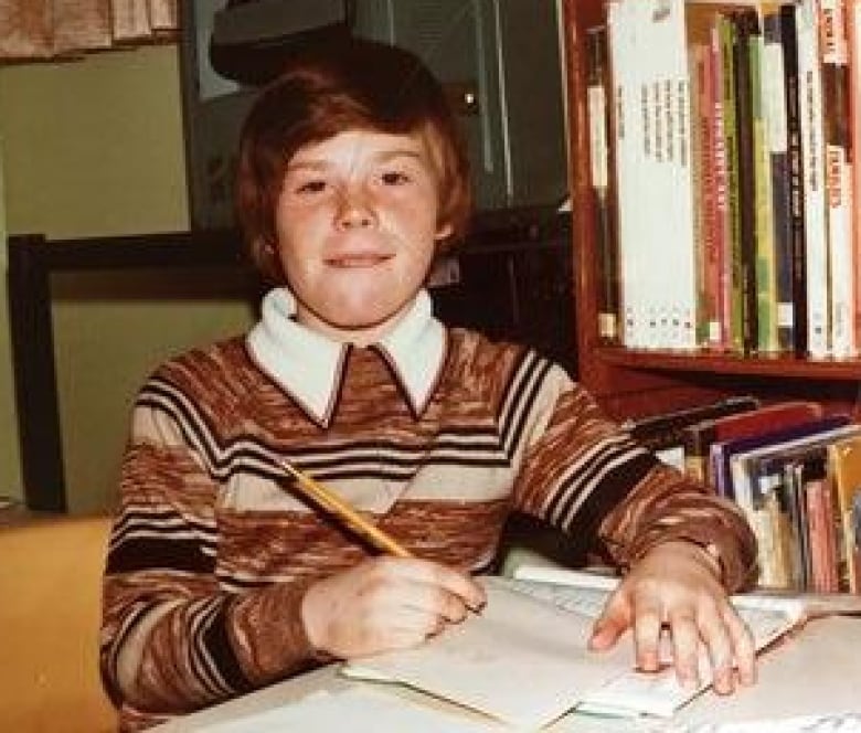 A clearly old photograph shows a young boy sitting in a library with a book and pencil.