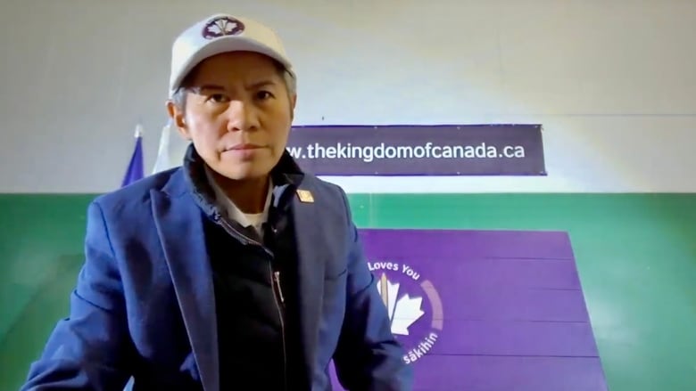 A woman wearing a white ball cap looks at the camera. A banner on the wall behind her says 'thekingdomofcanada.ca.'