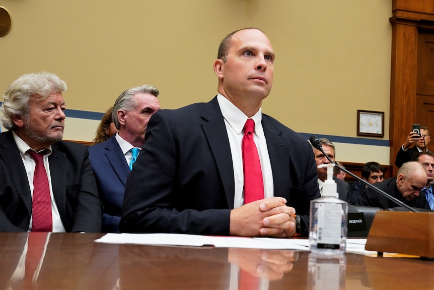 A man in a suit and red tie sits at a table, looking composed