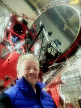 a man in a blue vest stands in a large room before a large red piece of equipment and a large dark circular disk towering over him.