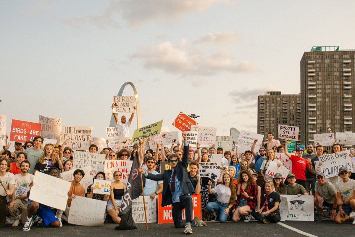 A sizable crowd in front of the Gateway Arch, holding "Birds Aren't Real" signs.