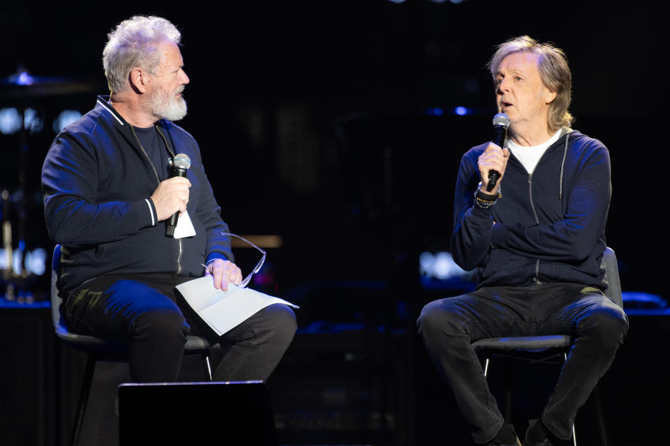 Paul McCartney and another man are seated on stage, holding microphones, having a conversation at an event