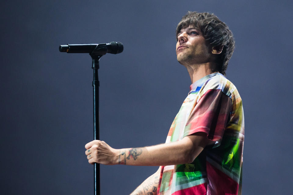 Louis Tomlinson stands on stage holding a microphone. He is wearing a colorful, loose-fitting T-shirt with tattoos visible on his arms