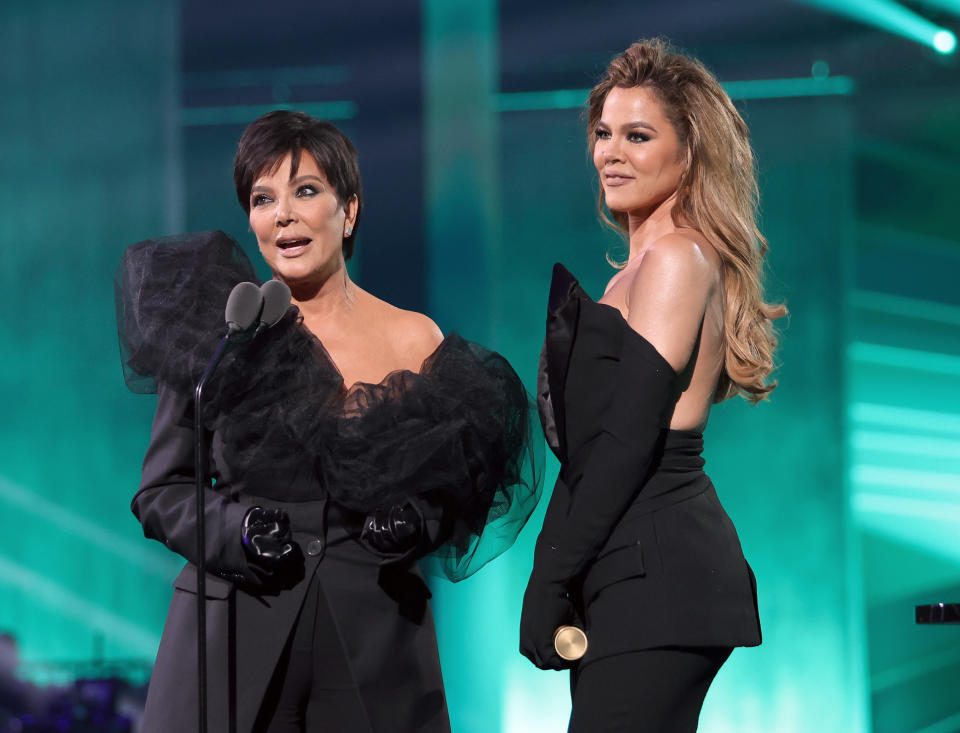 Kris Jenner and Khloé Kardashian on stage at an event. Kris wears an elegant black outfit with sheer ruffles, and Khloé in a chic black strapless gown