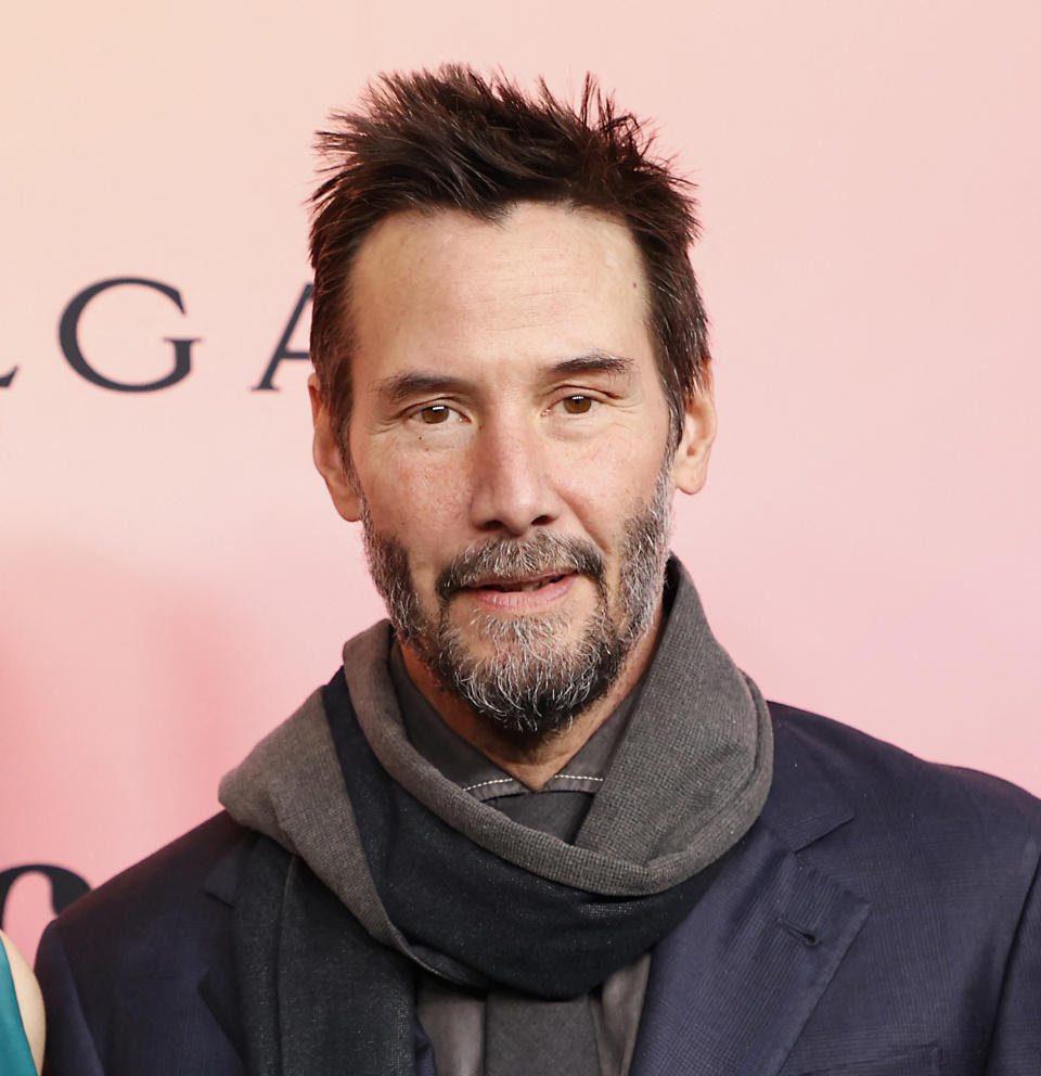 Keanu Reeves stands on a red carpet wearing a dark blazer and a gray scarf, with a trimmed beard and slightly messy hair