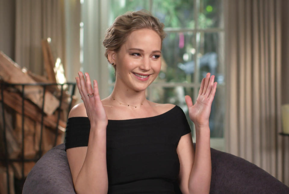 Jennifer Lawrence, smiling, with hands raised, sits indoors wearing an off-shoulder black dress