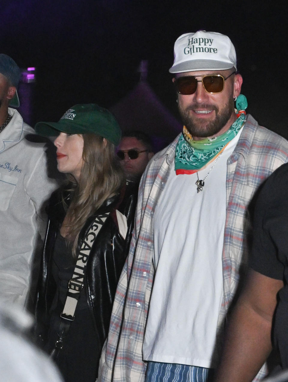 Taylor Swift and Travis Kelce at an event, with Taylor in casual attire and Travis wearing a plaid shirt and bandana. Travis wears a "Happy Gilmore" cap