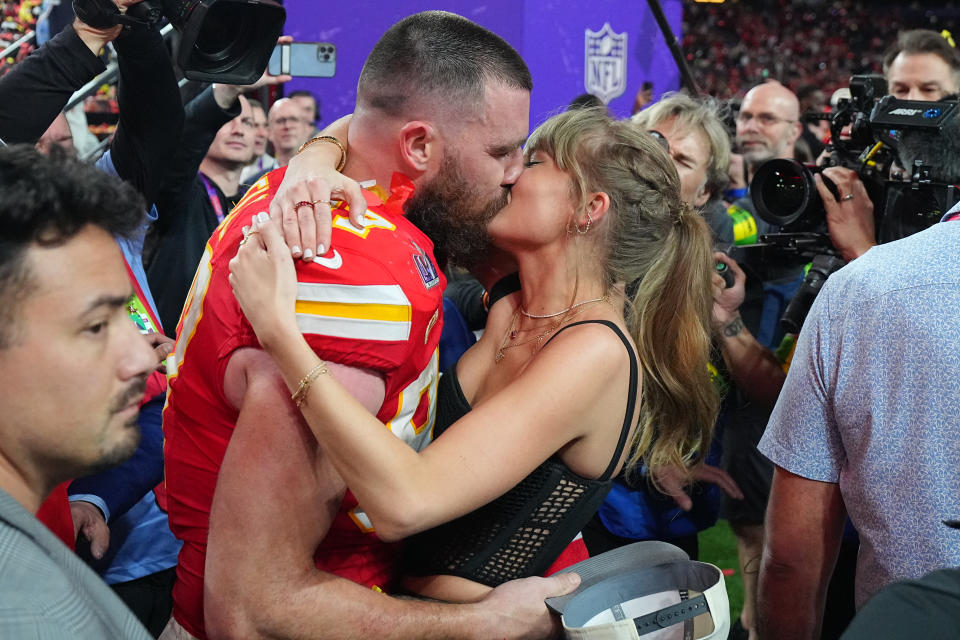 Travis Kelce, in a football uniform, and Taylor Swift, in a stylish dress, share a kiss surrounded by people and photographers at a crowded event