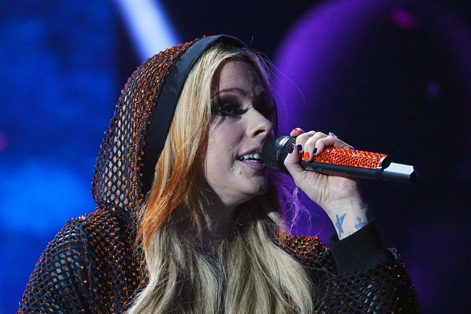 Avril Lavigne sings into a microphone on stage, wearing a hooded, sparkly mesh outfit