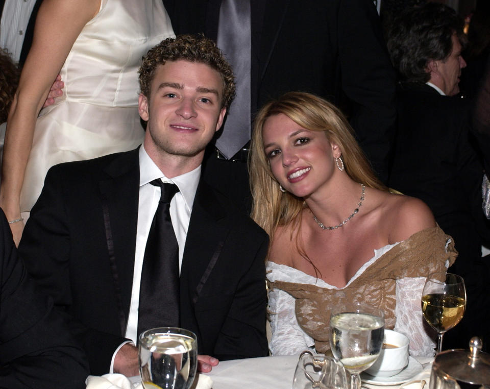 Justin Timberlake and Britney Spears sitting at a dinner event, both smiling at the camera. Justin is wearing a suit, and Britney is wearing an off-shoulder gown