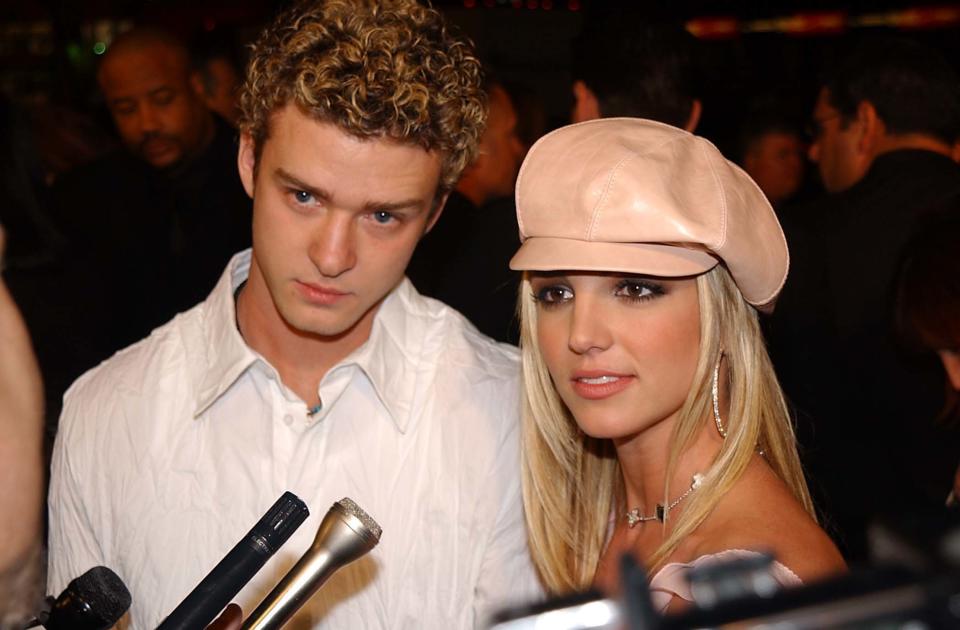 Justin Timberlake in a white shirt and Britney Spears in a light beret and shiny top with large earrings, surrounded by microphones at a media event