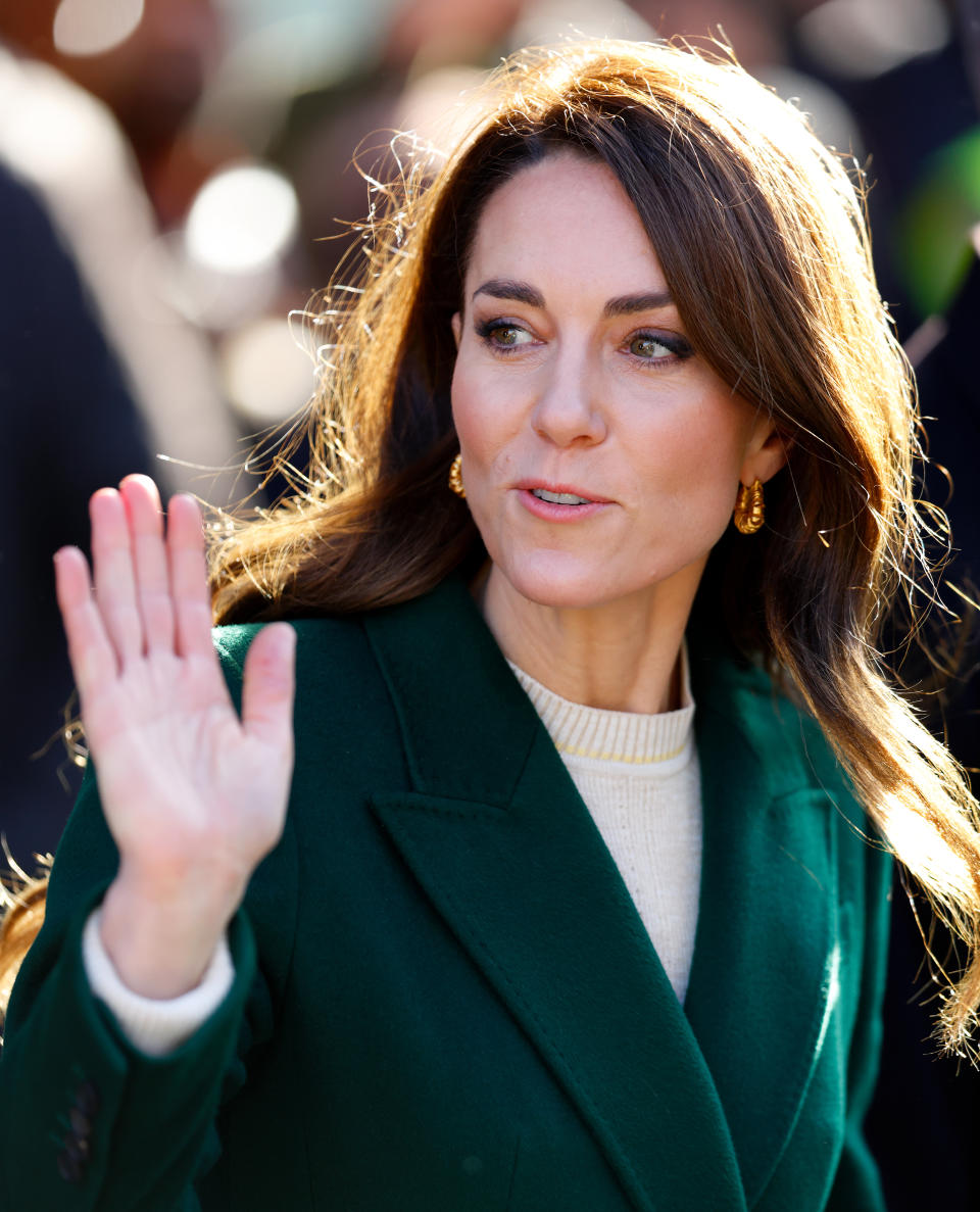 Kate Middleton waves and smiles while wearing a green coat and white top, with her hair down and gold earrings