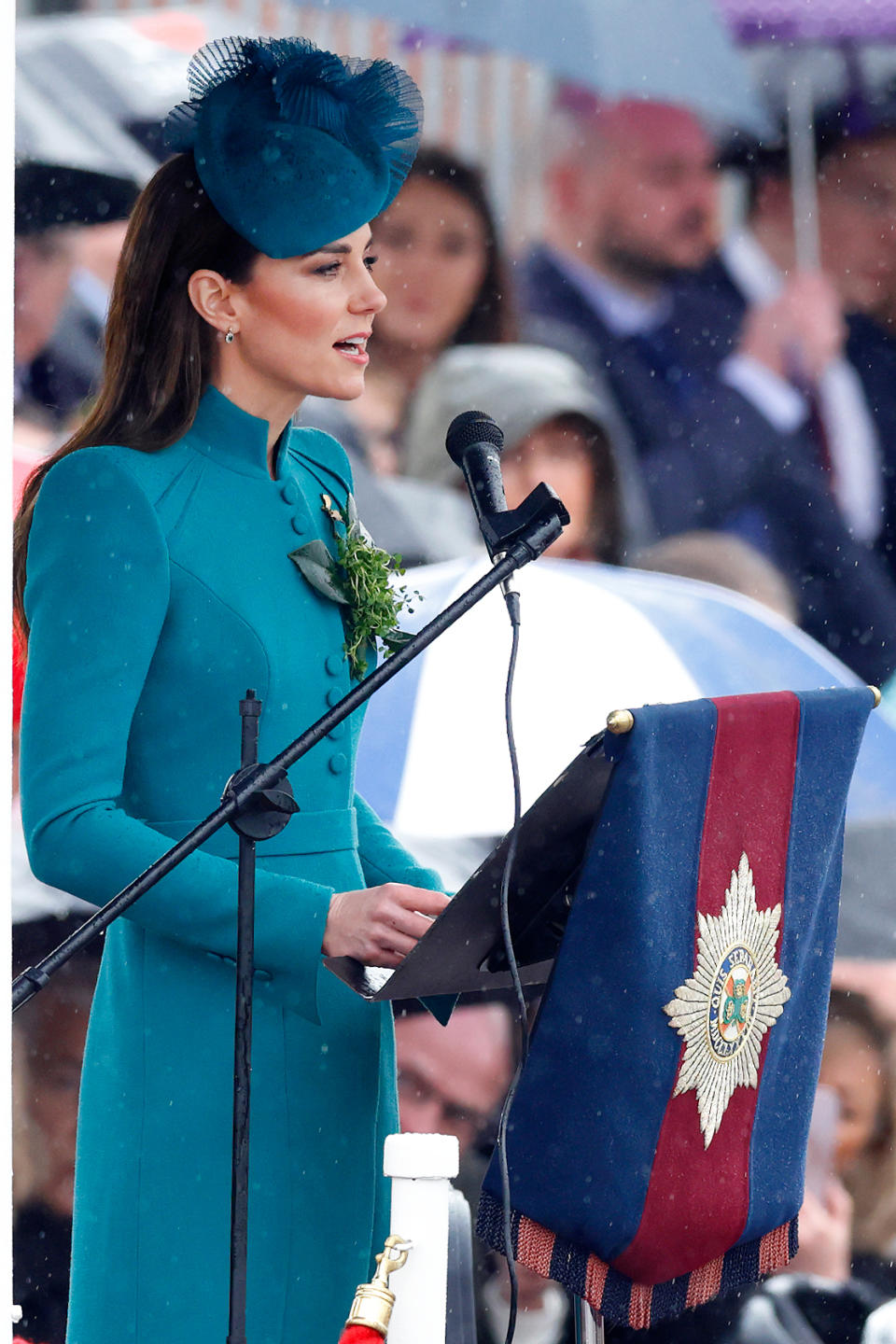 Kate Middleton giving a speech at an event, wearing a stylish formal dress with matching hat. People with umbrellas can be seen in the background due to rain