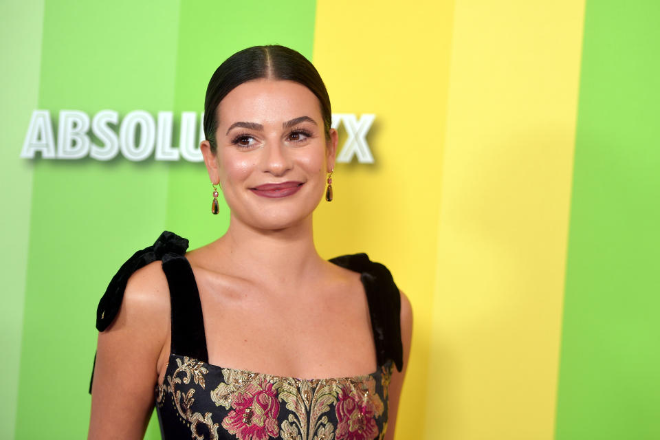 Lea Michele on a multicolored backdrop, wearing a floral embroidered dress with black velvet straps and statement earrings, smiling at the camera