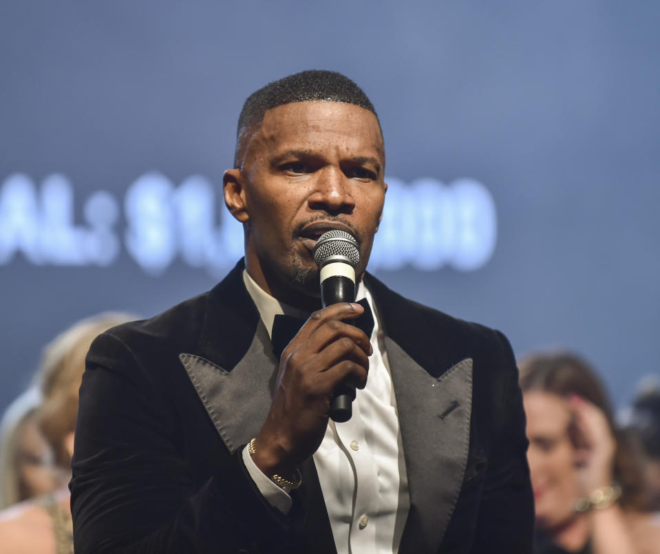 Jamie Foxx, wearing a black tuxedo with a white shirt, holds a microphone while speaking at an event with people visible in the background