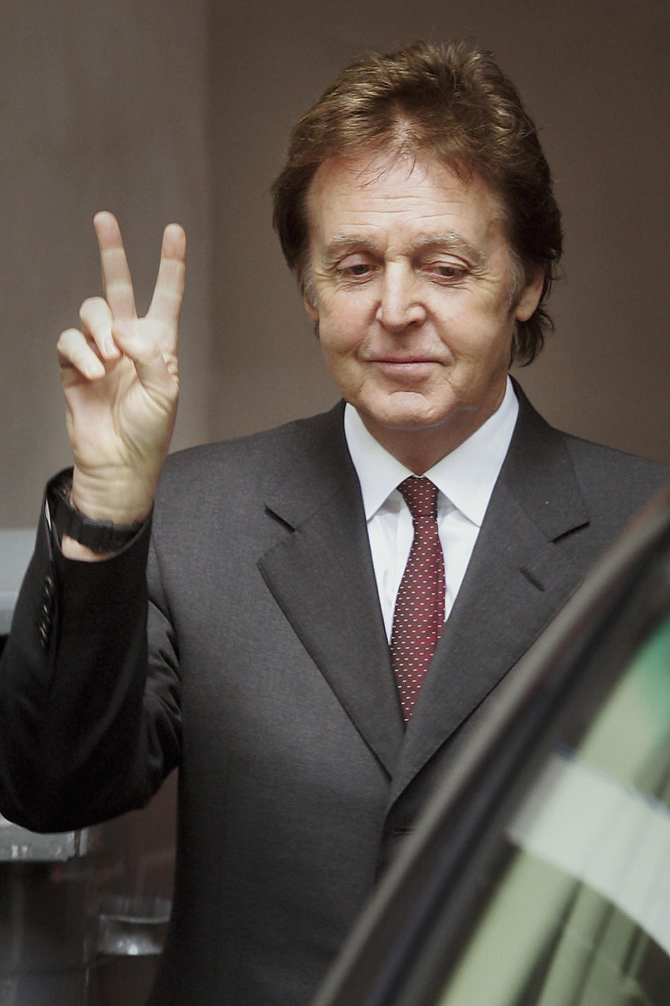 Paul McCartney in a suit and tie, making a peace sign with his right hand, while smiling slightly