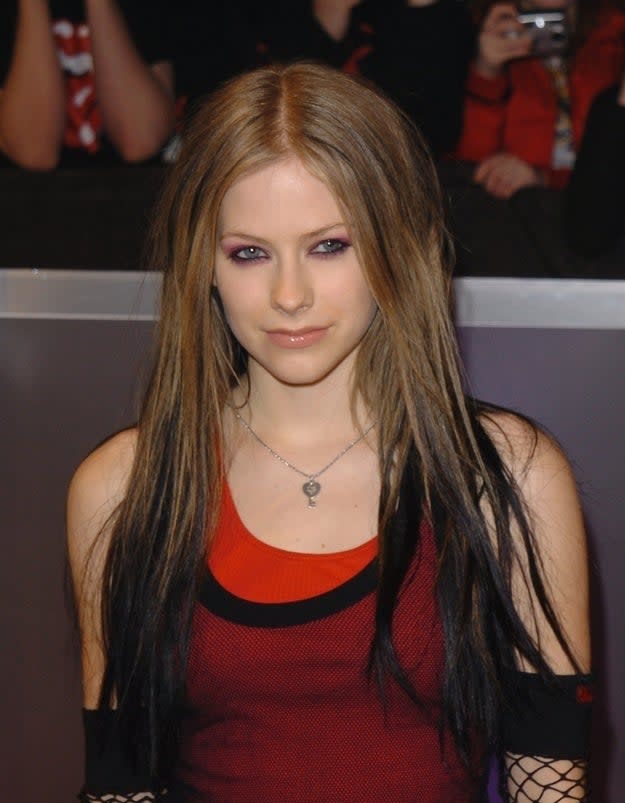 Avril Lavigne at an event, wearing a black and red sleeveless top with fishnet arm warmers. She has long, straight hair with dark streaks and a silver necklace